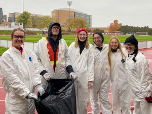 Five people in white protective suits stand in front of a soccer field holding a bag used for trash audits.