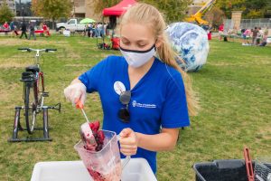 Morgan barlin wearing a blue shirt and white mask, pouring fruit into a blender cup in front of a grassy background
