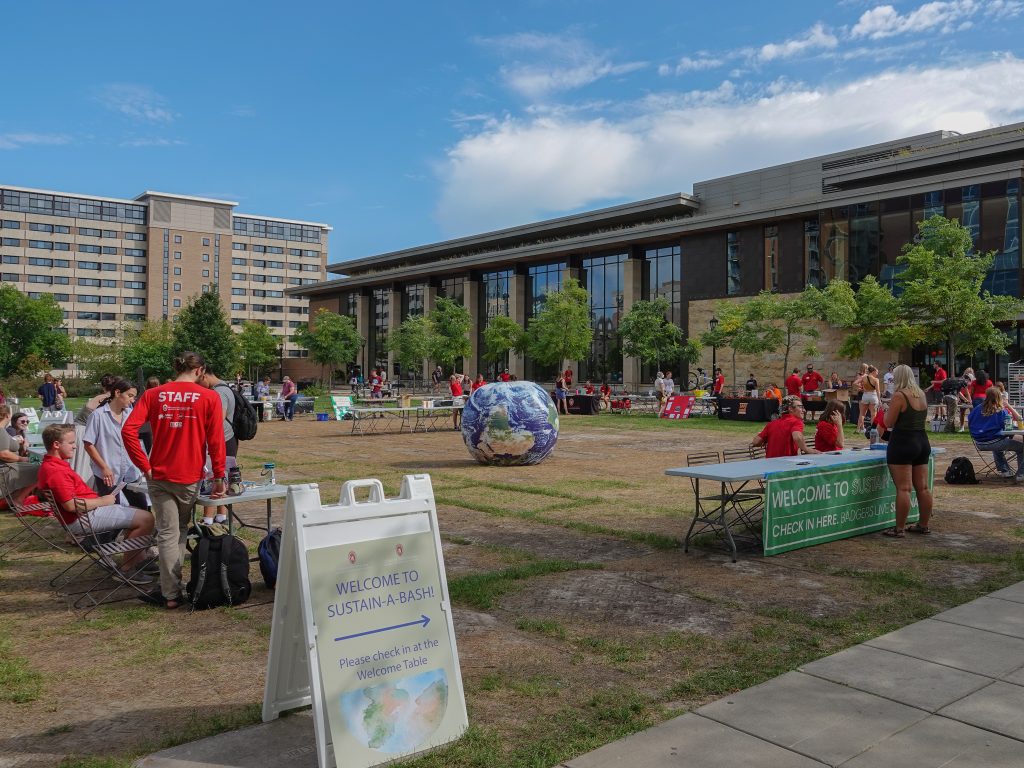 Field among campus buildings with signs saying "Sustain-a-Bash" and students mingling at tables