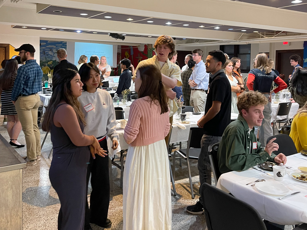 People sitting and standing, conversing, in a room with white covered tables.