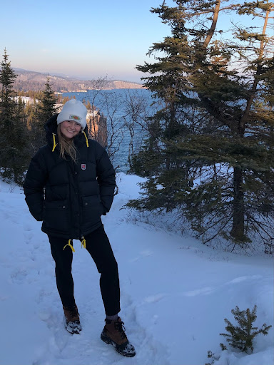 Cora Klemme wearing black pants and a coat and white hat standing in a snowy landscape. 