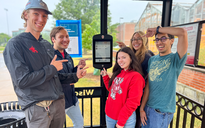 Students celebrate a project milestone, pointing to one of the new solar-powered displays at a bus shelter on Walnut Street. Their active engagement in the project showcases the campus as a living laboratory for sustainable transportation.