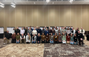 Conference attendees pose for a group photo in front of poster boards in the conference ballroom.