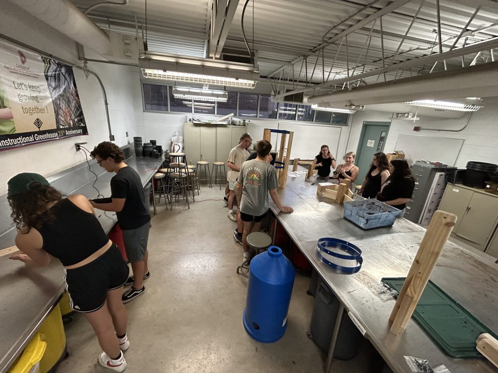 The image shows 8 students in a classroom working on building wooden structures for the aquaponics system.