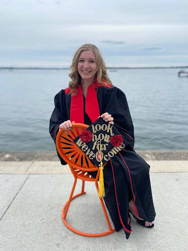 Mackenzie, donning her black graduation robes with red collar, smiles while seated on a terrace chair in front of Lake Mendota.