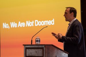 A man stands at a lectern in front of a slide that says "No, We Are Not Doomed"
