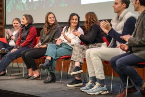 Seven panelists sitting on a stage, smiling and clapping.