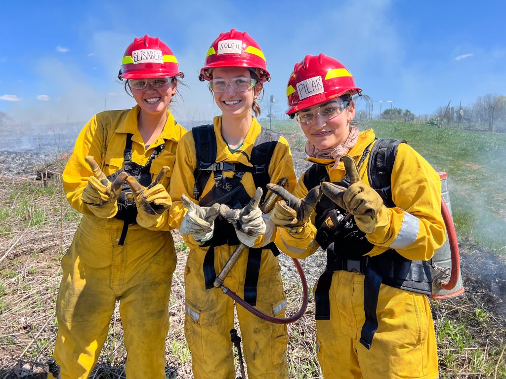 Three students standing in a prairie wearing protective Nomex clothing. 