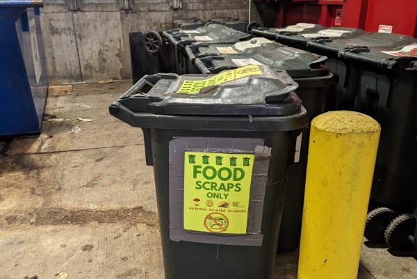In a facilityu0027s loading dock and trash collection area, a photo showing a line of rolling bins with the one at the end displaying a bright yellow sign that reads u0022Food Scraps Only.u0022