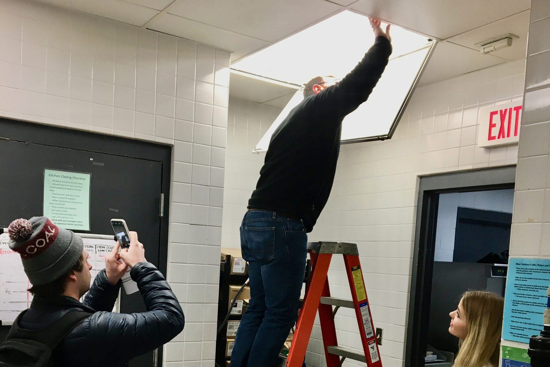 A staff member on a ladder installs new ceiling lighting while two students observe.