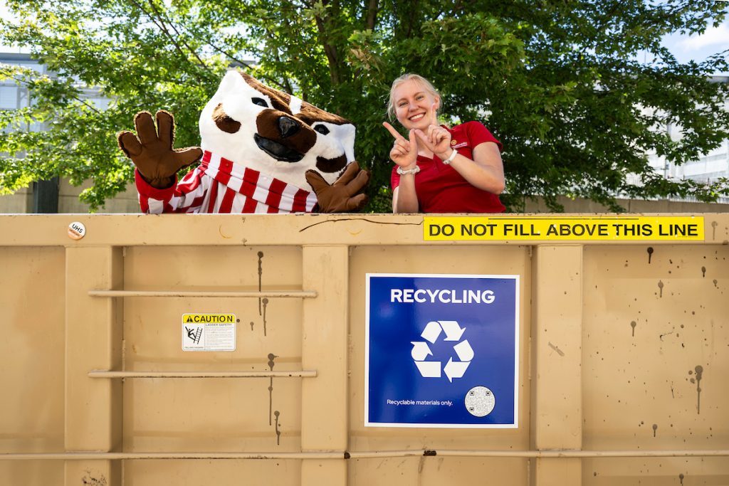 UW–Madison mascot Bucky Badger stands in a recycling dumpster next to a student wearing a red polo.