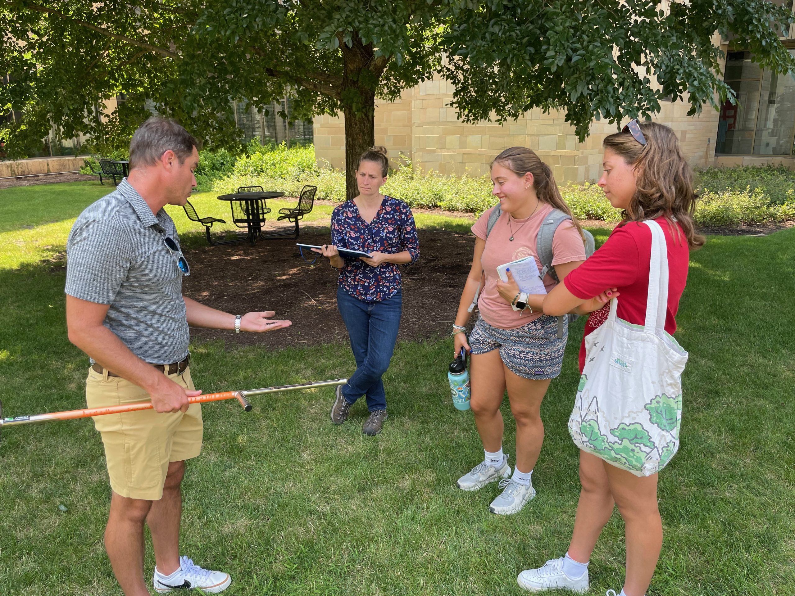 A group of four students and staff inspect a soil sample at a grassy site.
