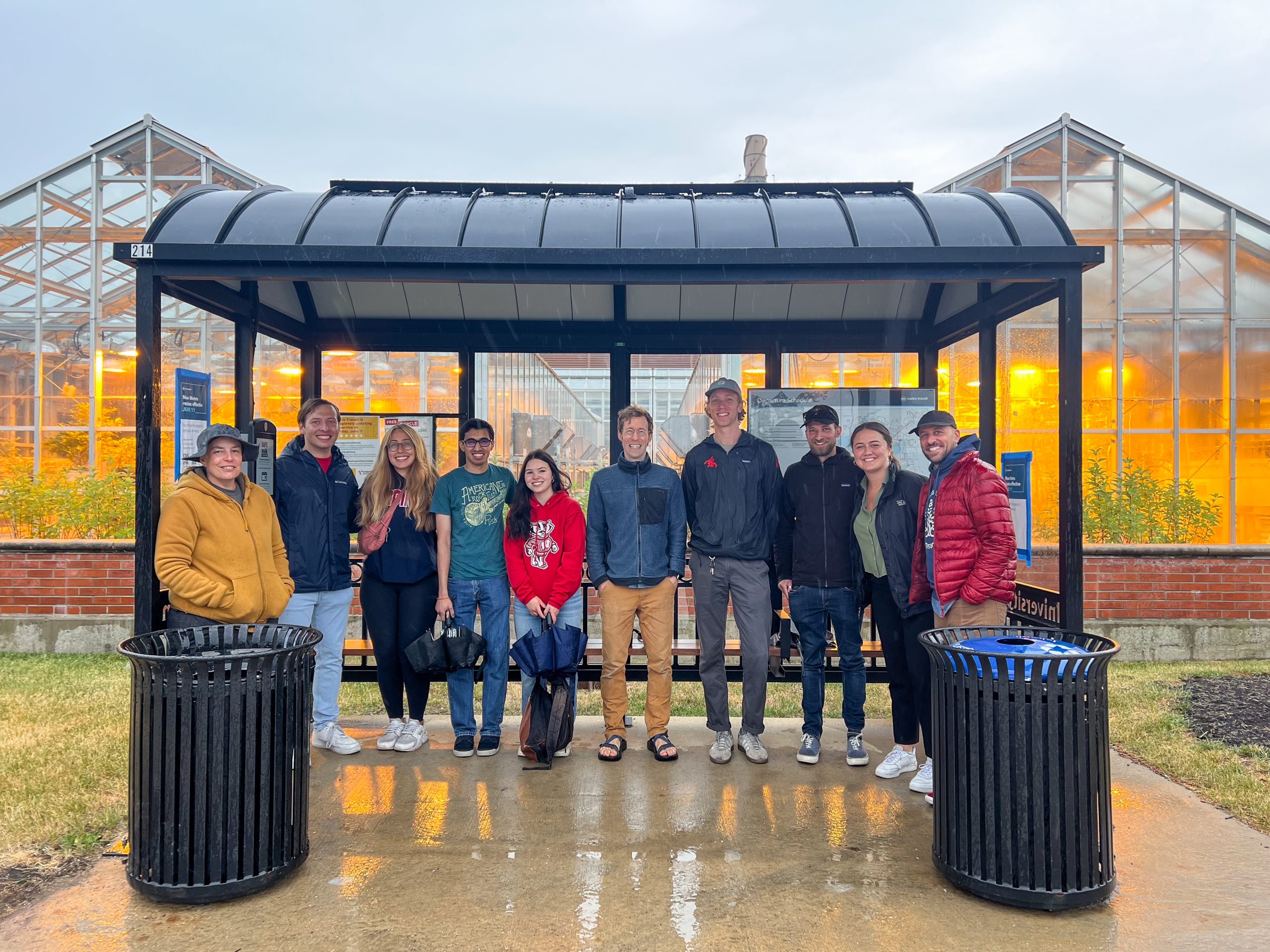 ten project partners gather inside a campus bus shelter that has curved solar panels installed on the roof