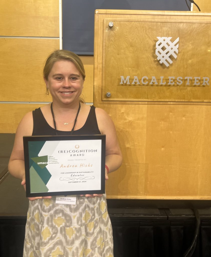 Andrea Hicks smiling with a plaque in her hands standing in front of a podium.