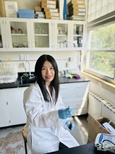 Mohan Qin working in a lab, wearing a white coat and blue gloves.