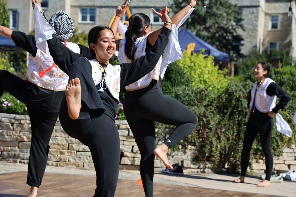 Indian dancers move with their hands up and one foot in the air, holding scarves
