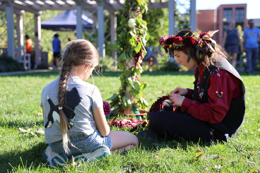 A student and child sit in the grass making flower crowns