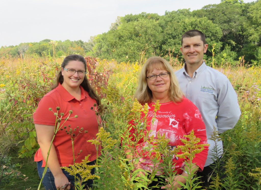 Bryn Scriver, Laura Wyatt, and Adam Gundlach smile while standing in a field of yellow flowers with trees in the background.