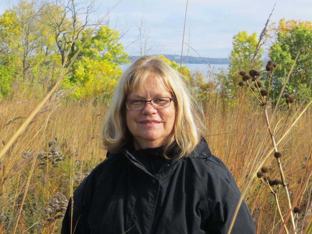 Laura Wyatt wearing a black jacket smiling amidst tall prairie plants with a lake and yellow trees in the background.