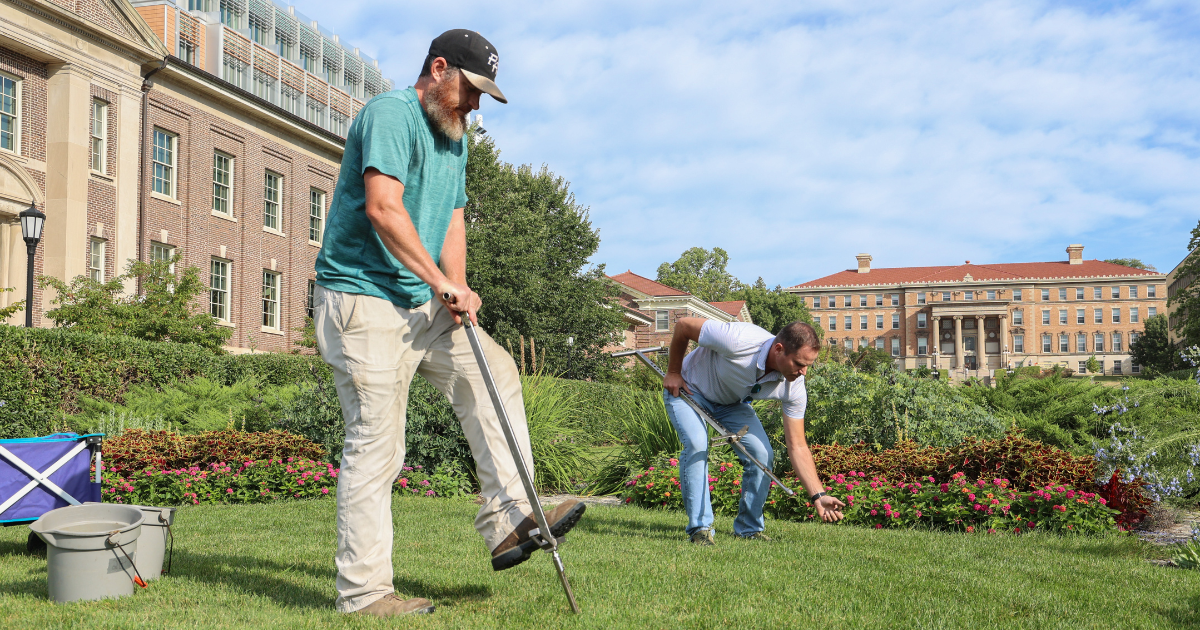 Two men collect soil samples on a lawn in front of a large building at UW–Madison, using tools and standing among flower beds on a sunny day.