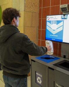 A student in a black shirt interacting with an LCD screen sitting on top of a row of waste and recycling bins.