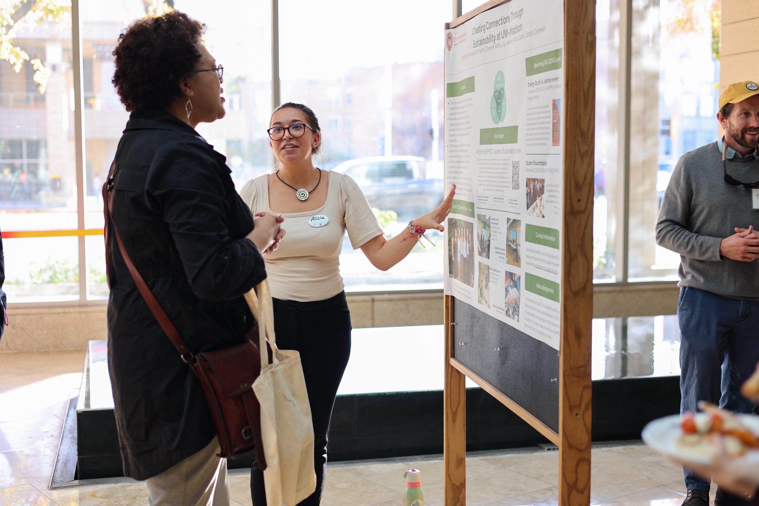 Two people stand next to a poster board in front of a large wall of windows.