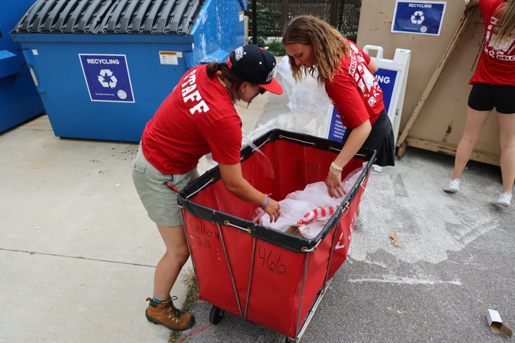 Two students look into a red Housing rolling bin filled with plastic bags.
