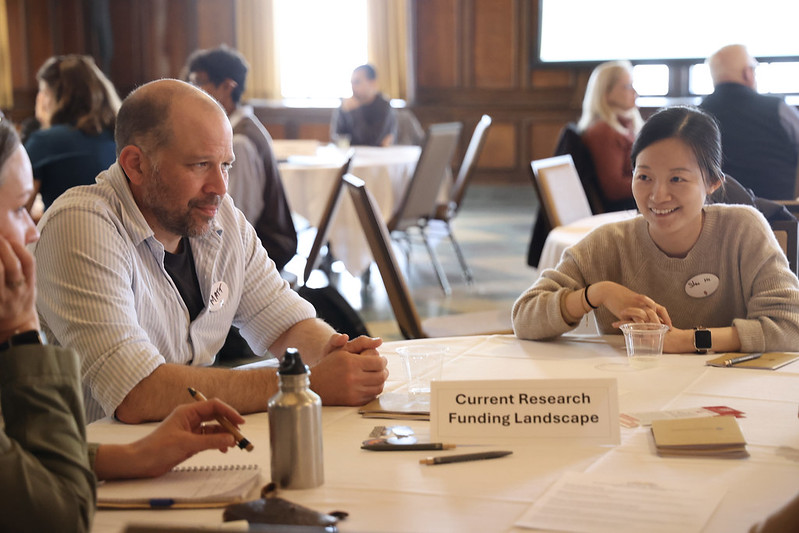 Matt Ginder-Vogel sits at a round table with other participants during a sustainability research networking event. A table sign reads “Current Research Funding Landscape,” and attendees are engaged in conversation with notebooks, pens, and drinks on the table.