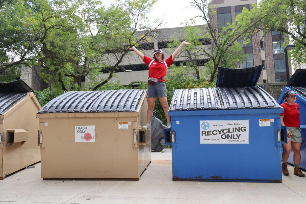 Student stands between trash and recycling dumpsters with her hands in the air.