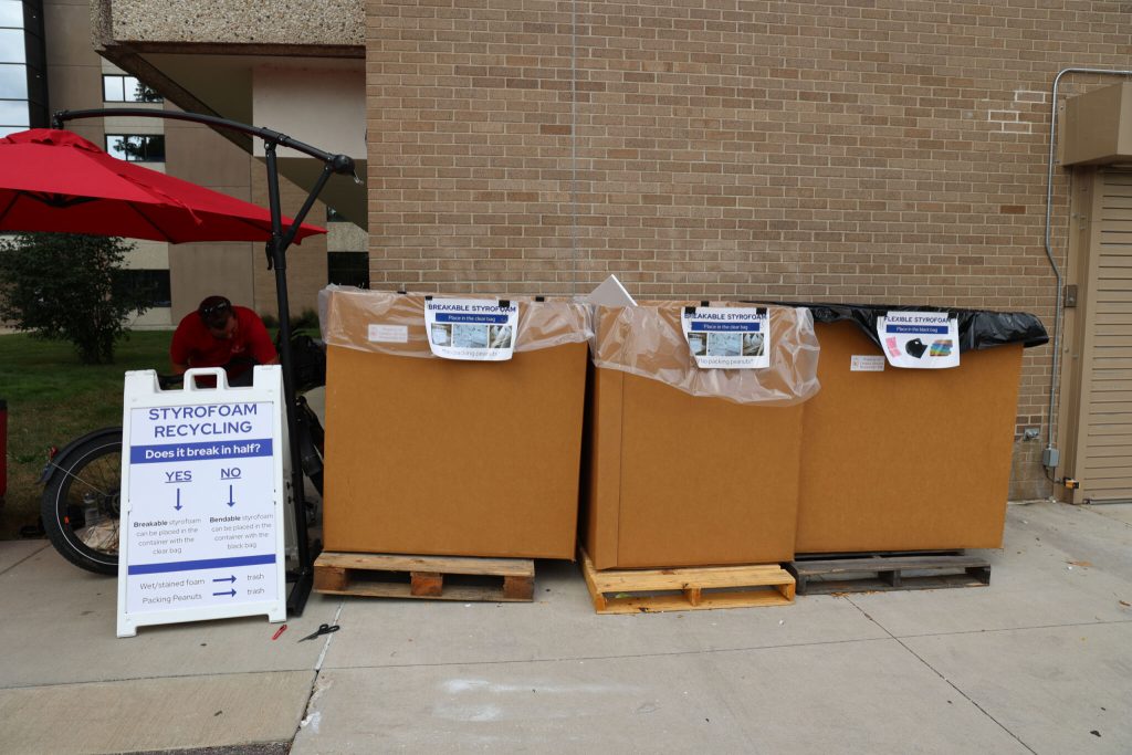 Three brown bins with labels for various types of stryfoam and a standing sign with stryofoam recycling instructions.
