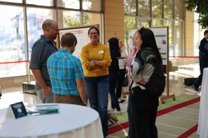 A group of people stand and talk near a table at a bright indoor event space during the Sustainability Symposium. Some hold coffee cups and wear name tags, while posters and large windows are visible in the background.