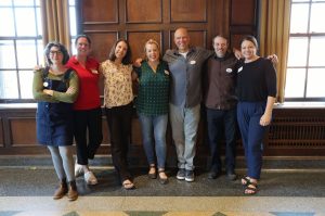 Seven members of the Sustainability Research Hub team stand together, smiling for a group photo in a wood-paneled room.