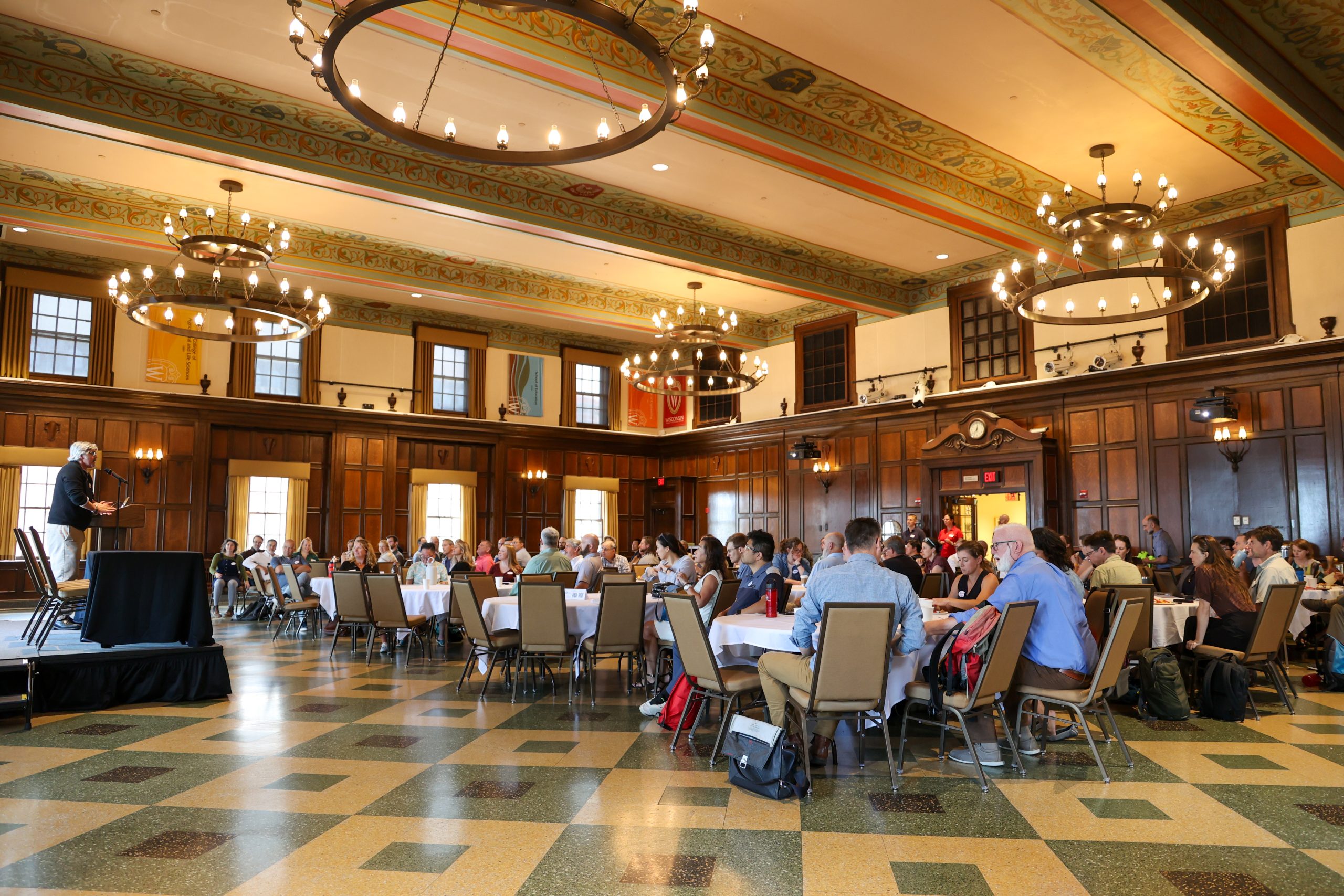 A large group of attendees sits at round tables in a grand hall with chandeliers and wood paneling, listening to Paul Robbins speak at a podium during the fall 2024 Sustainability Research Visioning Event.