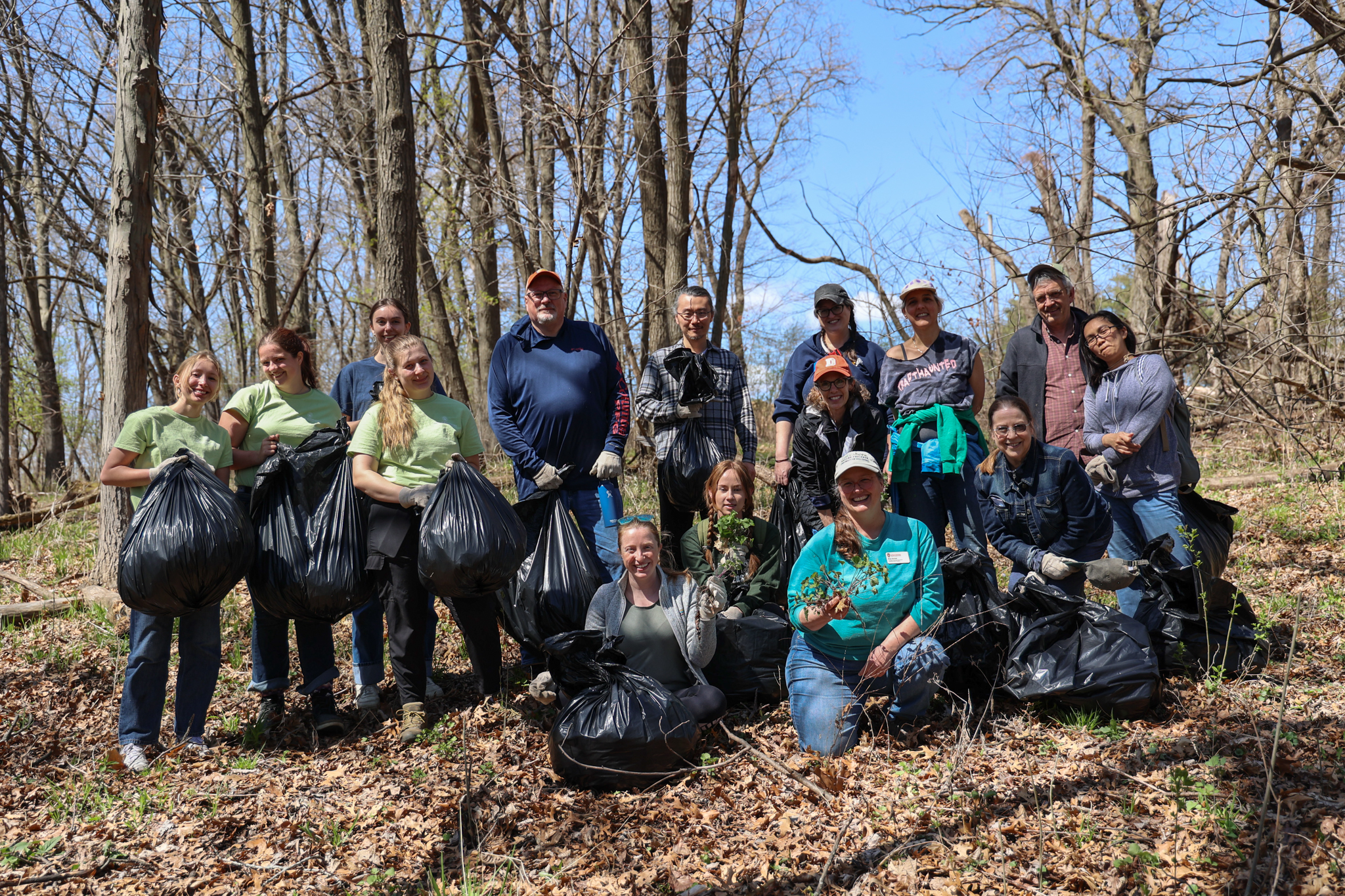 A group of about fifteen people pose together in the woods holding black garbage bags and plants after a volunteer restoration work day in the Lakeshore Nature Preserve.