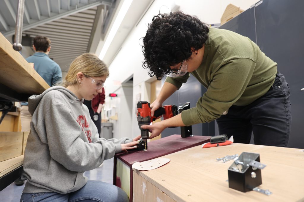 two students wearing safety glasses drill hardware into a piece of wood