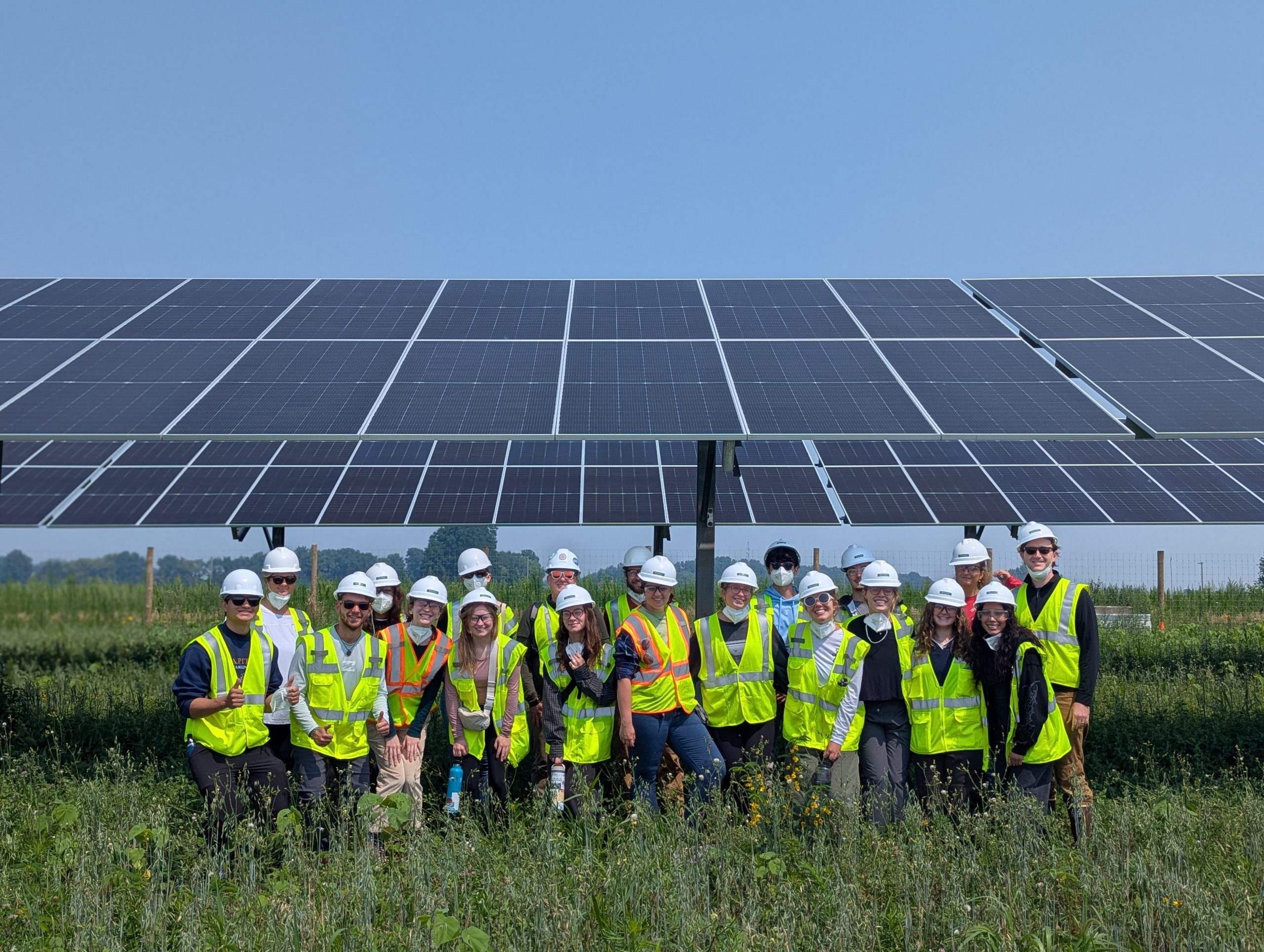 A group of about twenty people wearing bright yellow safety vests and white hard hats stand together in front of a large solar panel array in a grassy field.