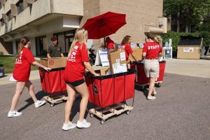 Students pushing red move in cards filled with boxes. The students are wearing read shirts that read "Staff" on the back. They are outside of a residence hall in the waste station area pushing the carts toward the dumpsters. A red umbrella is in the background covering student workers from the sun. 