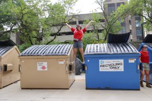 Student worker stands with hands in the air between a garbage dumpster and recycling dumpster during University Housing Move-In