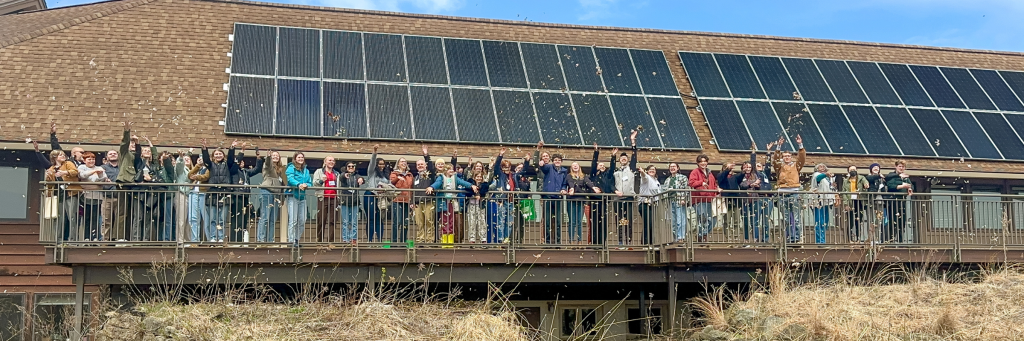 A large group tosses pale magnolia tepals into the air in celebration, with newly installed rooftop solar panels behind them at the UW–Madison Arboretum.