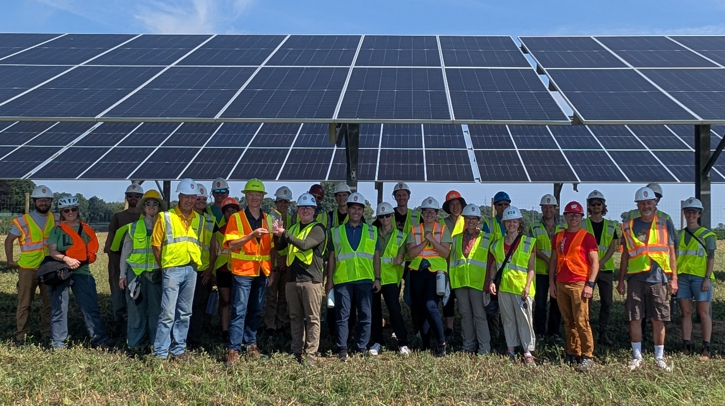 A large tour group wearing safety vests and hard hats stand smiling next to a large solar array.