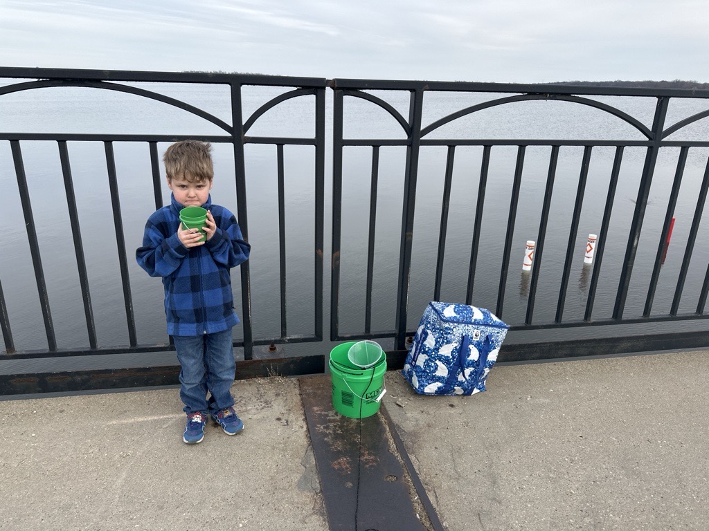 A small child standing by the lake with a bucket on the ground.