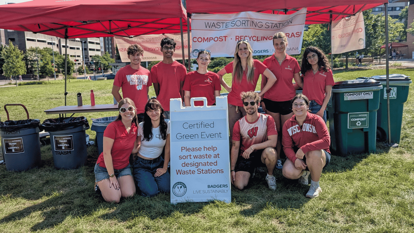 A group of people in red shirts stands behind a waste sorting station with recycling bins. A sign reads "Certified Green Event. Please help sort waste at designated Waste Stations."