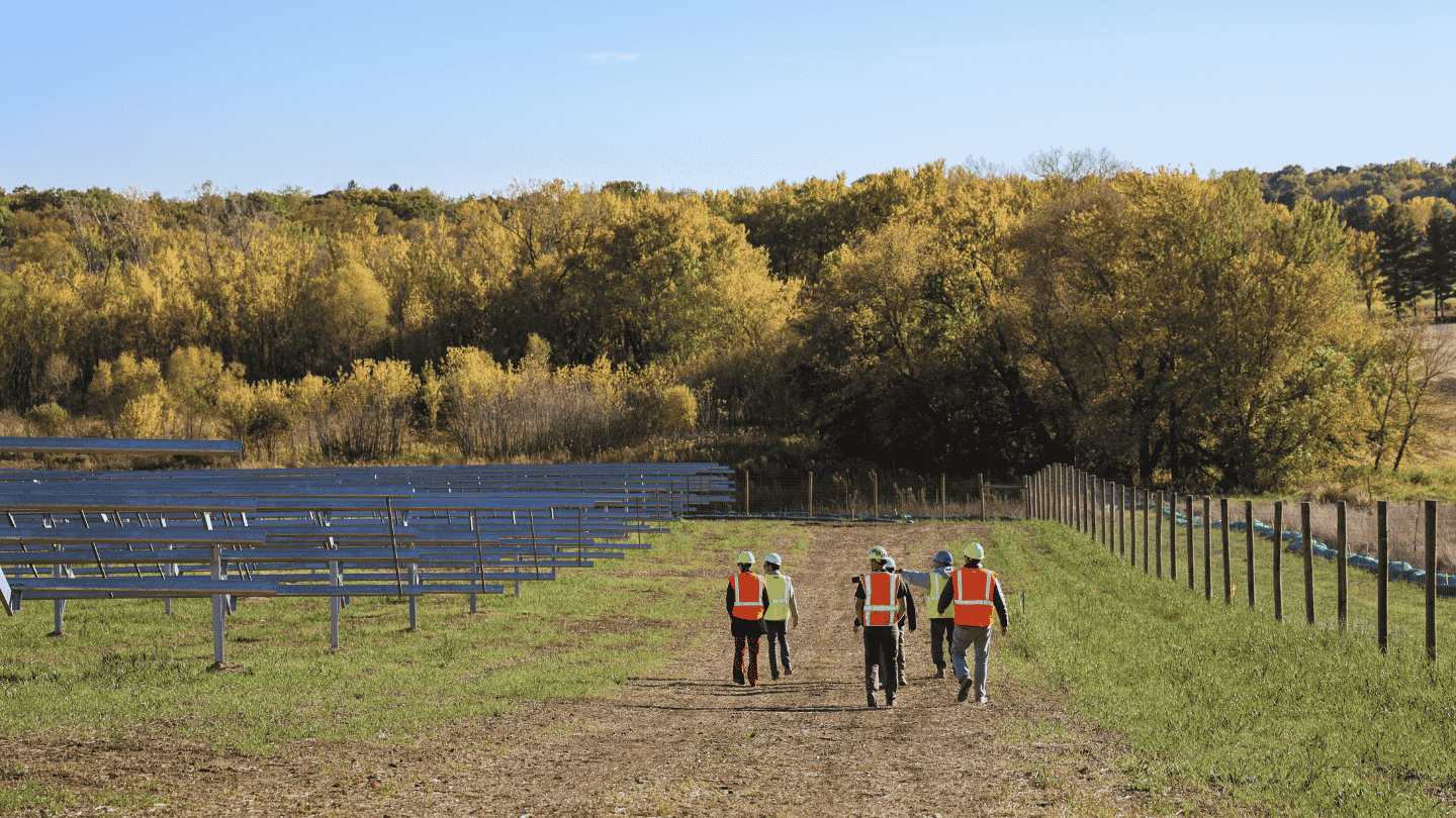 A group of people in high-visibility vests walk on a dirt path next to rows of solar panel racking, with a forest in the background.