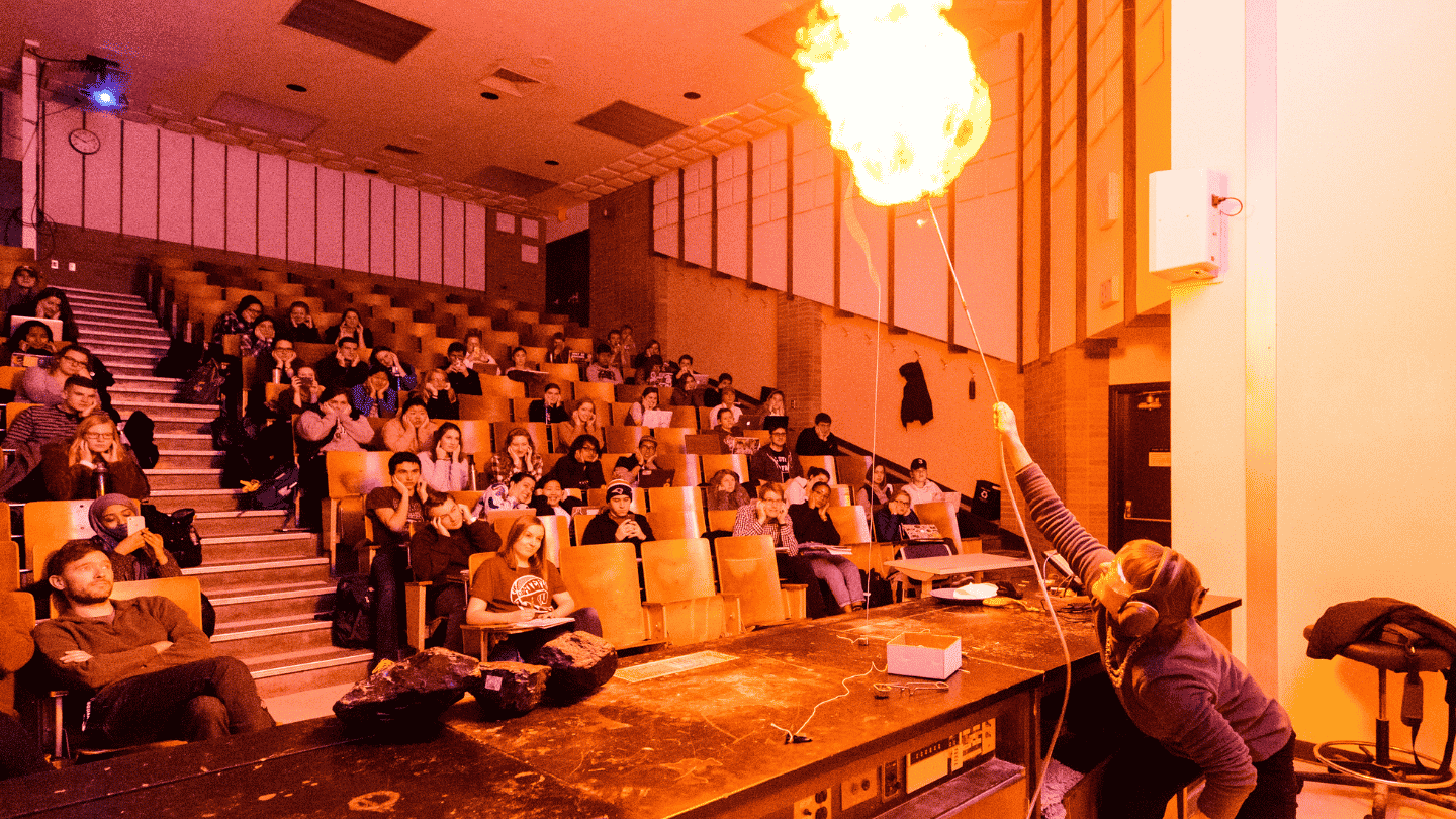 A woman stands at the front of a full lecture hall conducting a science experiment.