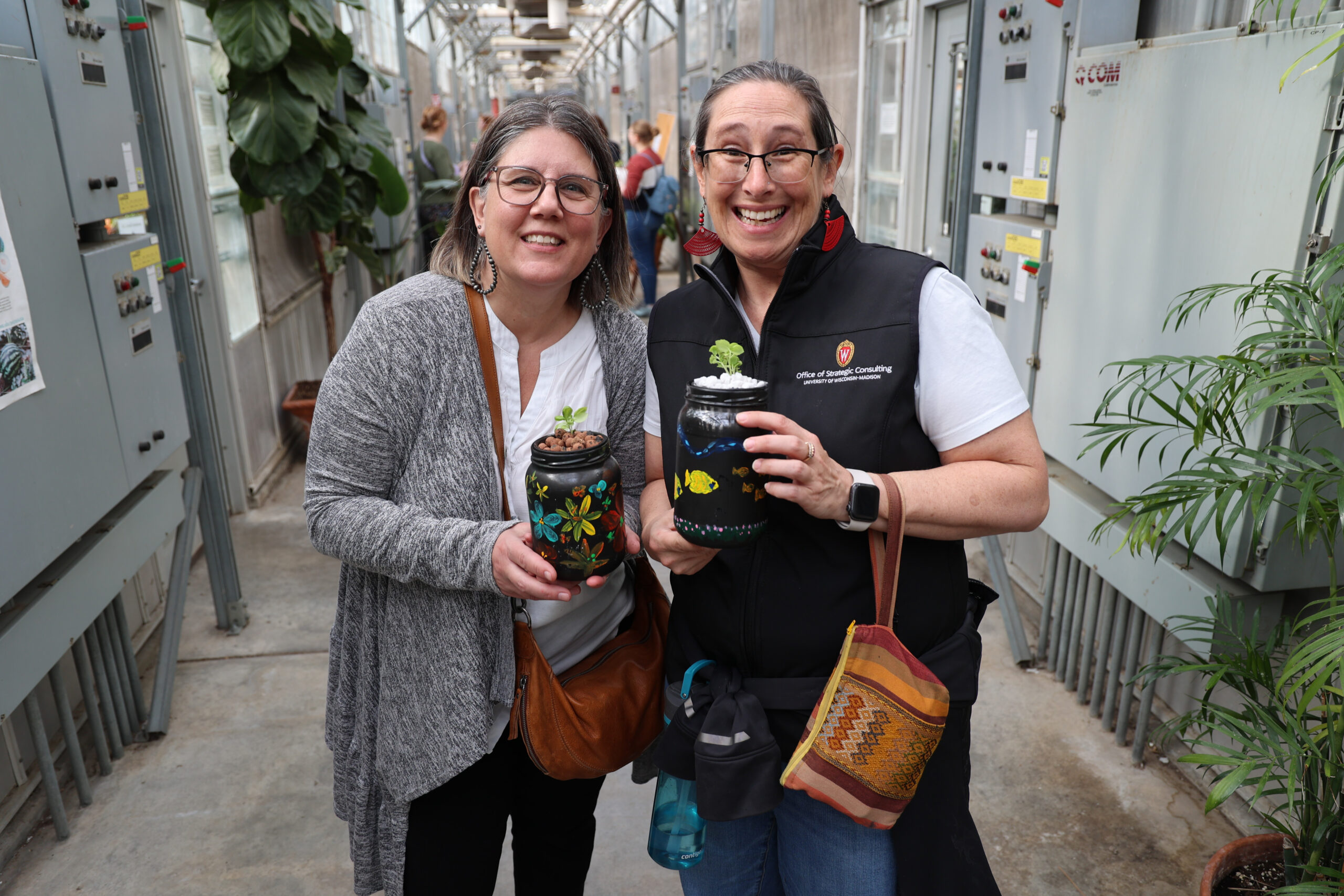 Two staff members stand side-by-side in a greenhouse, smiling and holding hand-painted mini aquaponics jars.