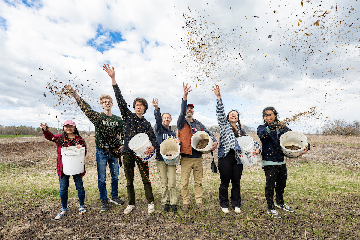 seven people stand side-by-side, holding buckets and casting wildflower and prairie seeds into the air