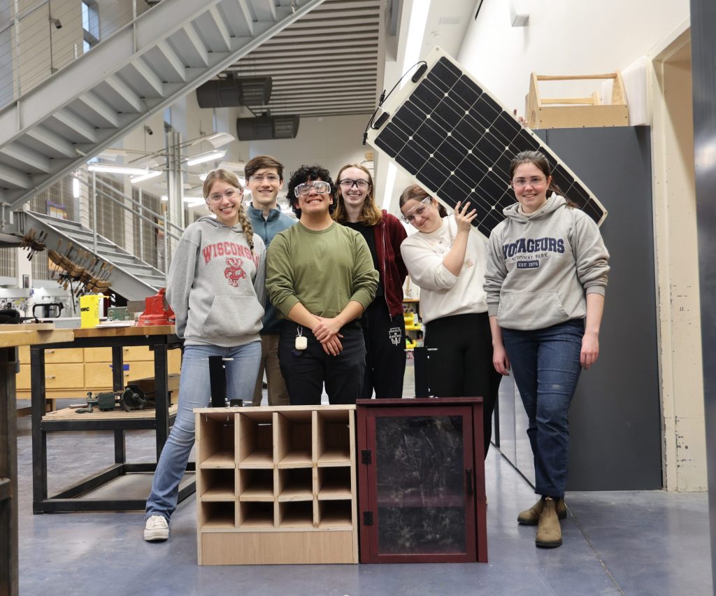 Students gather around a little free library, smiling and holding up a solar panel.