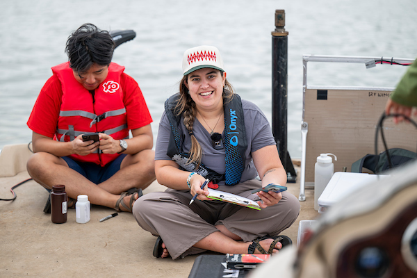 Two students in life vests sit on a boat, smiling, and holding clipboards.