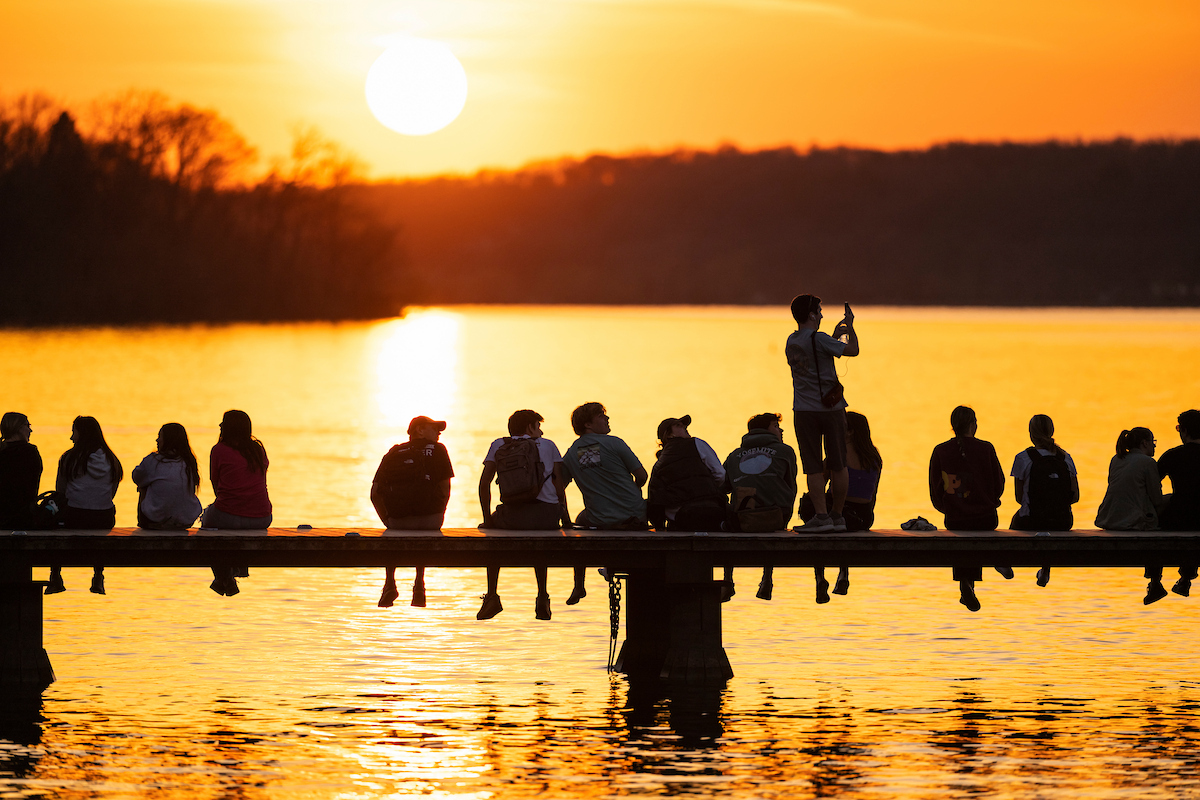 Students sit along a pier silhouetted against a bright orange sunset over Lake Mendota, while one person stands to take a photo.