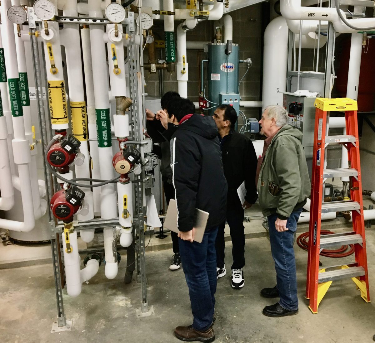 Four men stand in a utility room examining pipes